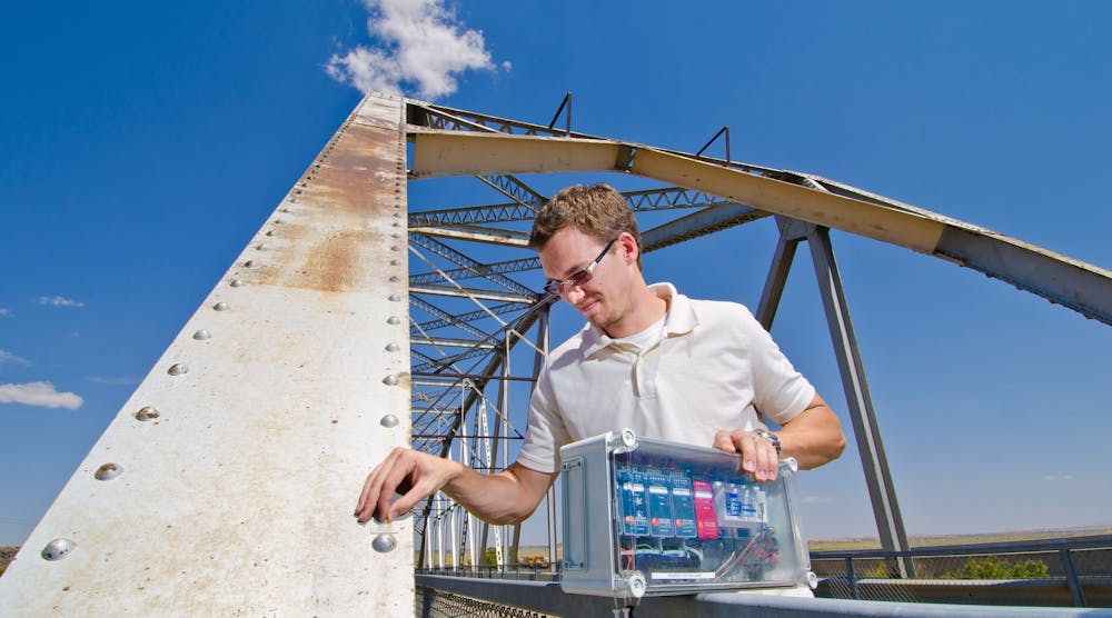 Sandia National Laboratories mechanical engineer Stephen Neidigk positions a comparative vacuum monitoring sensor on a bridge. In his other hand is the controller that periodically checks the sensor and a wireless transmitter to autonomously alert maintenance engineers if it detects a crack.