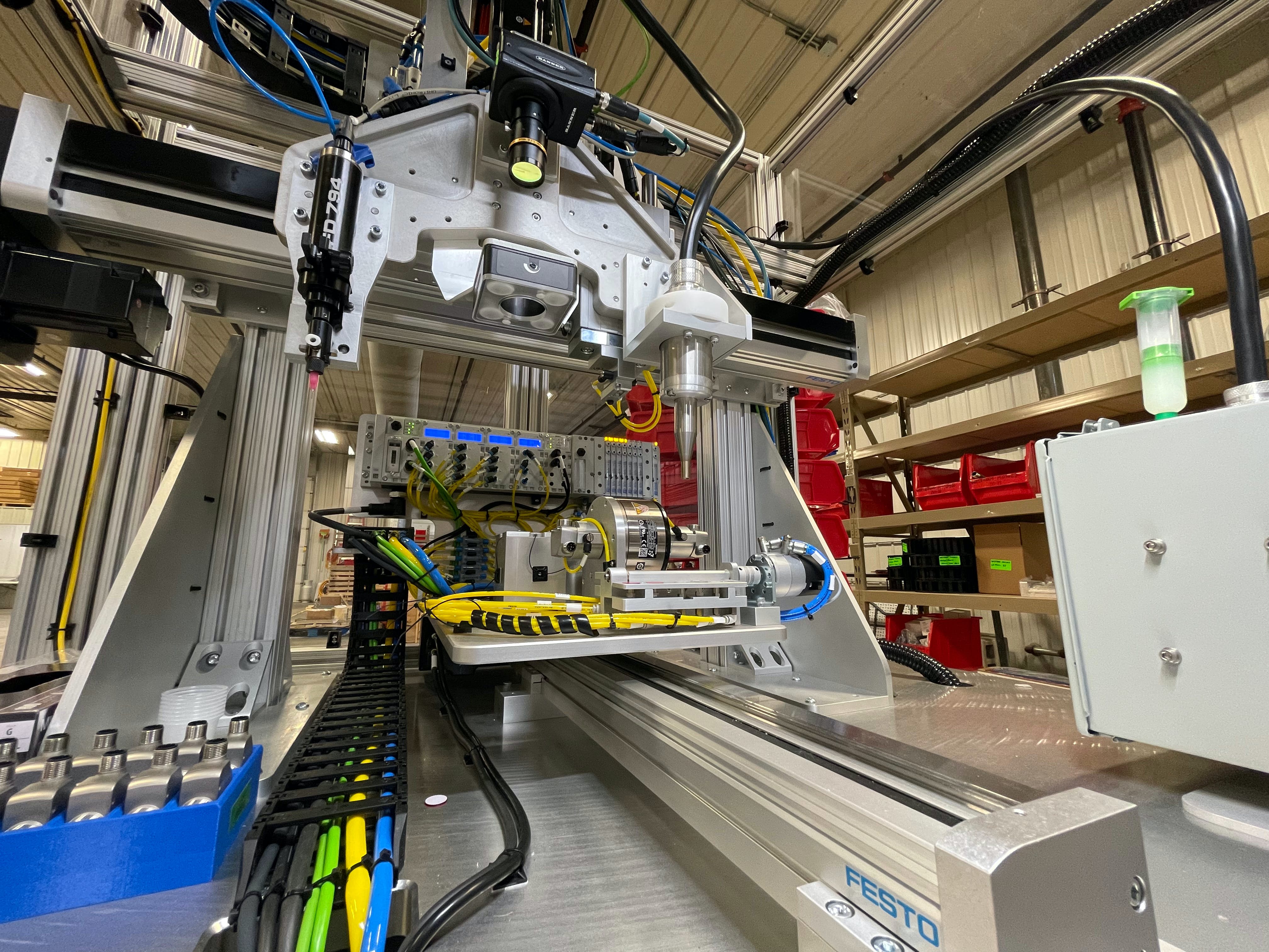 A closeup view of the Y axis, center, and the Z and X axes, above. On the Z axis from left to right are the Nordson adhesive applicator, Banner vision system and plasma gas applicator. The pick-and-place guided actuator is mounted behind the plasma applicator and is not easily distinguished. A box of die-cast housings is visible on the lower left.